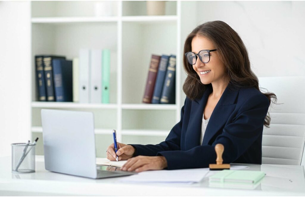 Selbstbewusste Geschäftsfrau mit Brille stempelt an einem hellen Schreibtisch ein Dokument und arbeitet am Laptop, um Multilingua Translation Service zu symbolisieren